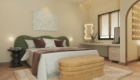 Giường ngủ trung tâm với đầu giường bọc nệm màu xanh rêu uốn lượn, kết hợp cùng ánh sáng vàng dịu nhẹ từ đèn thả và tranh nghệ thuật tối giản phía trên